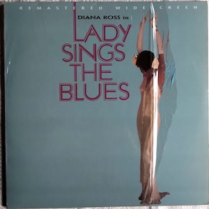 May include: A blue and white VHS tape cover with the text "DIANA ROSS in LADY SINGS THE BLUES" in pink lettering. A woman in a white dress is standing with her arms raised.