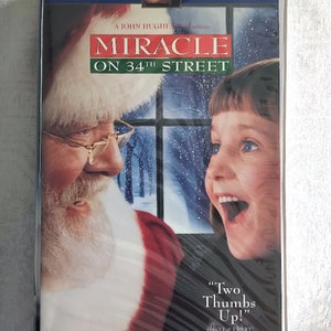 May include: A VHS tape cover for the movie "Miracle on 34th Street". The cover features a close-up of Santa Claus and a young girl looking at each other. The text "Two Thumbs Up!" is printed on the bottom right corner of the cover.
