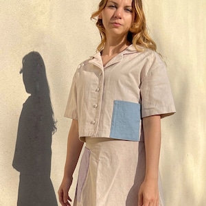 Può includere: Una donna indossa un completo in lino beige in due pezzi con una tasca a toppa blu sulla parte superiore. Indossa sandali dorati con borchie.