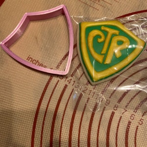May include: A pink plastic cookie cutter in the shape of a shield sits next to a decorated cookie. The cookie is shield-shaped with a green center and gold border, featuring the letters "CTR".