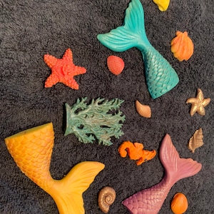 May include: Assortment of colorful mermaid tail soaps, seashells, starfish, and seahorse soaps. The soaps are arranged on a dark gray textured surface.