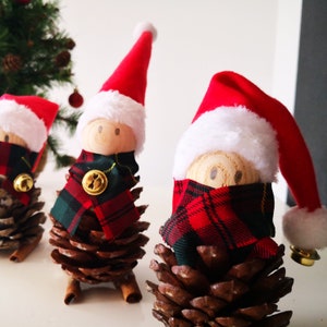 Può includere: Decorazioni natalizie fatte a mano con pigne, teste di bambole di legno e sciarpe a quadri rossi e verdi. Ogni ornamento ha un cappello da Babbo Natale rosso con finiture bianche e una piccola campana dorata. Un piccolo albero di Natale è sullo sfondo.