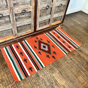 May include: A Southwestern-style rug with a geometric pattern in shades of orange, red, black, white, and green. The rug has a fringe border.