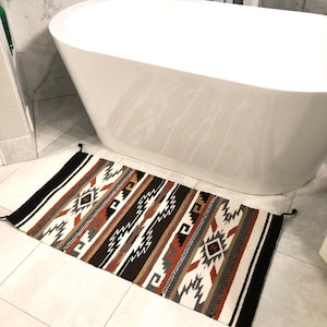 May include: A white bathroom rug with a black, brown, and orange geometric pattern. The rug is in front of a white bathtub.
