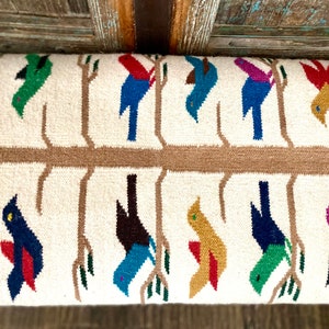 Op de afbeelding: Een wit geweven kussen met een bruin boomtakontwerp en kleurrijke vogels. De vogels zijn in verschillende kleuren, waaronder rood, blauw, groen, geel en paars.