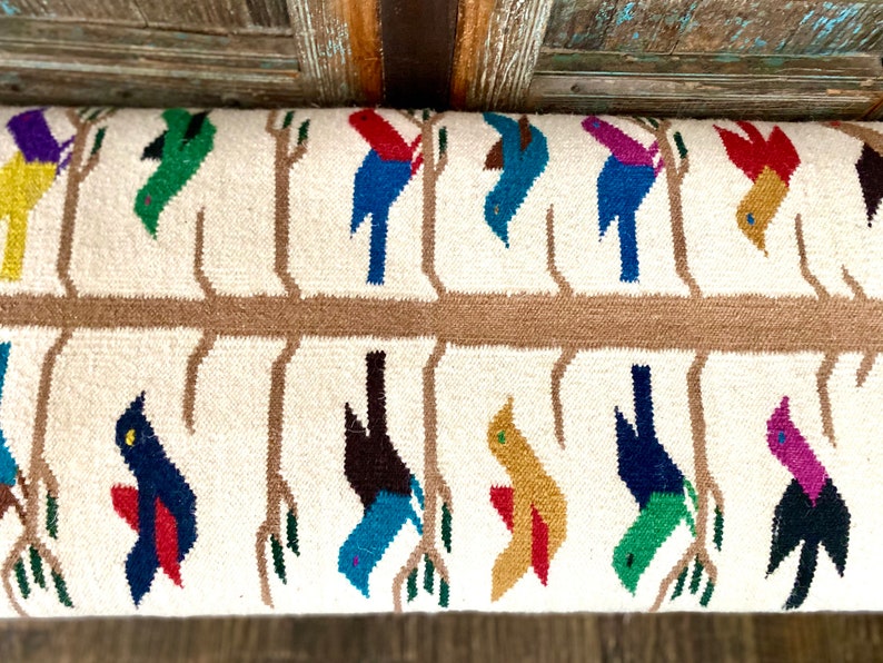 Op de afbeelding: Een wit geweven kussen met een bruin boomtakontwerp en kleurrijke vogels. De vogels zijn in verschillende kleuren, waaronder rood, blauw, groen, geel en paars.