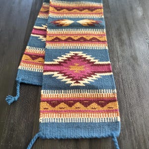 May include: A woven textile scarf in shades of blue, pink, gold, and burgundy. The scarf features geometric patterns and tassels at the ends. The design includes diamond and zigzag motifs, creating a Southwestern or tribal aesthetic.
