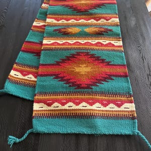 May include: A woven table runner with a Southwestern design. The runner features a teal background with geometric patterns in red, orange, and cream. The ends have teal tassels. The runner is approximately 60 inches long.