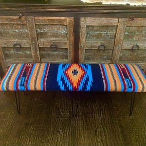 May include: A blue, orange, and red patterned bench with black metal hairpin legs. The bench is in front of a wooden cabinet with three doors.