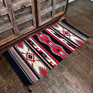 May include: A rectangular, woven rug with a Southwestern design. The rug features a black background with red, white, and gray geometric patterns. The rug has tassels on each end and is placed on a wooden floor.