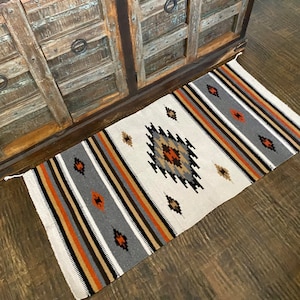 May include: A white, gray, black, and orange woven rug with a geometric pattern. The rug has a diamond shape in the center with stripes on the sides.