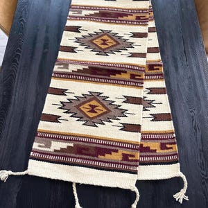 May include: A white table runner with a geometric pattern in shades of brown, gold, and burgundy. The runner has fringe on the ends.