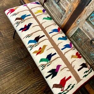 Op de afbeelding: Een witte bank met een geweven ontwerp van een boom met kleurrijke vogels. De bank heeft een bruin houten frame en zwarte metalen poten.