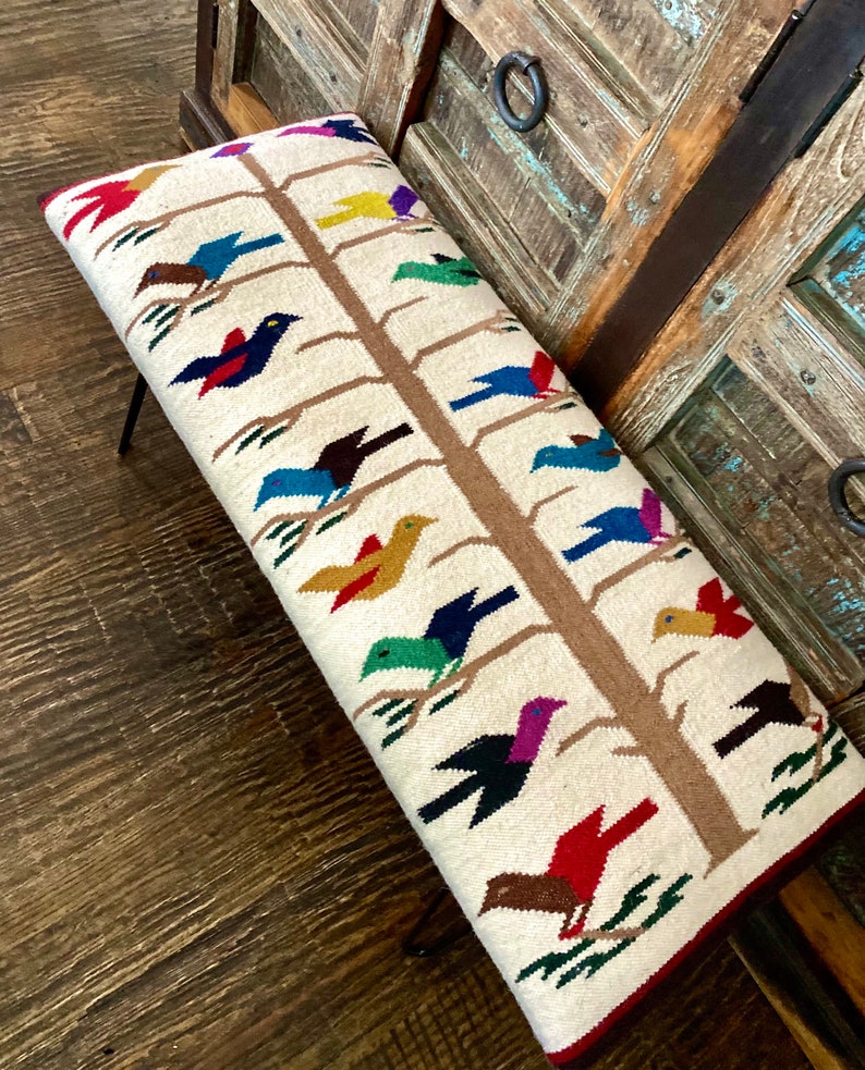 Op de afbeelding: Een witte bank met een geweven ontwerp van een boom met kleurrijke vogels. De bank heeft een bruin houten frame en zwarte metalen poten.