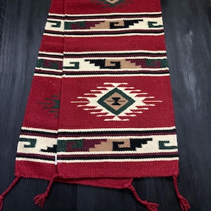 May include: A red woven textile with a geometric pattern in white, black, green, and tan. The textile has a diamond-shaped central design and tassels at the ends. The textile is likely a table runner or decorative scarf.