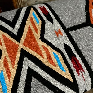 K&ouml;nnte beinhalten: Nahaufnahme eines grauen gewebten Textils mit geometrischem Muster. Das Design weist schwarze, beige, orange, blaue und rote Akzente auf. Das Textil hat ein strukturiertes Aussehen, was auf ein strapazierf&auml;higes Material hindeutet. Das Muster umfasst Zickzack- und Rautenformen.