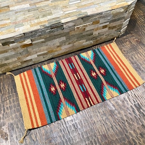 May include: A colorful woven rug with a geometric pattern in shades of red, orange, green, blue, and beige. The rug has a fringed edge and is made of natural fibers.