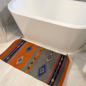 May include: A colorful, geometric patterned bath rug with tassels on the ends. The rug is orange, gray, blue, and green. The rug is in a bathroom with a white bathtub.