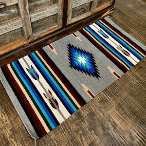 May include: A gray and white woven rug with blue, teal, and red stripes and a diamond pattern in the center. The rug is on a wooden floor.