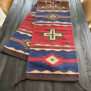 May include: A woven table runner with a Southwestern design. The runner features a repeating pattern of crosses, diamonds, and geometric shapes in red, blue, brown, and green. The runner is rectangular with tassels at the ends.