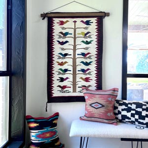 May include: A woven wall hanging with a tree design and colorful birds. The tapestry is cream-colored with a dark burgundy border. Decorative pillows in various patterns and colors are below the hanging.