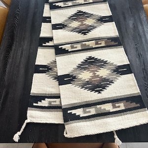 May include: Cream-colored woven table runner with a geometric diamond pattern in black, gray, and brown. The runner has a textured appearance and tassels at the ends. The design is reminiscent of Southwestern or tribal textiles.