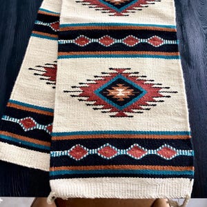 May include: Cream-colored woven table runner with a Southwestern design. The runner features geometric patterns in shades of red, turquoise, black, and brown. The design includes diamond and rectangular shapes.