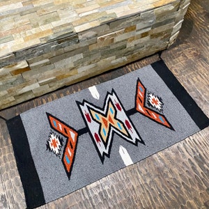 May include: A grey and black rug with a geometric design in orange, red, white, and blue. The design features a series of arrows pointing in different directions.