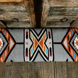 K&ouml;nnte beinhalten: Rechteckiges graues Kissen mit einem geometrischen Muster im S&uuml;dwesten. Das Design weist orange, schwarze, wei&szlig;e und blaue Akzente auf. Das Kissen liegt auf einer Holzoberfl&auml;che und unterstreicht seine dekorative Wirkung.