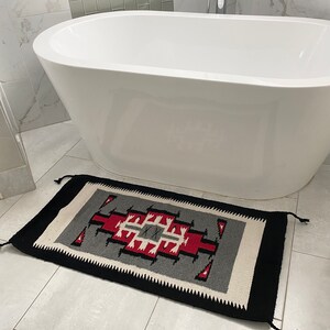 May include: Rectangular woven rug with a geometric design in red, gray, black, and white. The rug is placed on a tiled floor in front of a white bathtub. The rug has a black border and tassels on the ends.