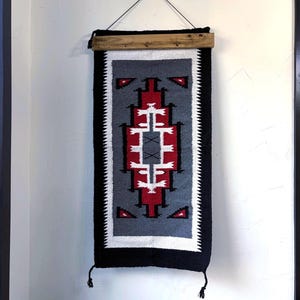 May include: A rectangular wall hanging with a geometric design in red, white, gray, and black. The textile is suspended from a wooden bar with a black cord. The design features triangles and a central pattern.