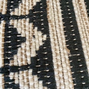 Puede incluir: Primer plano de una alfombra tejida con un patr&oacute;n negro y crema. La alfombra est&aacute; hecha de fibras naturales y tiene una superficie texturizada.