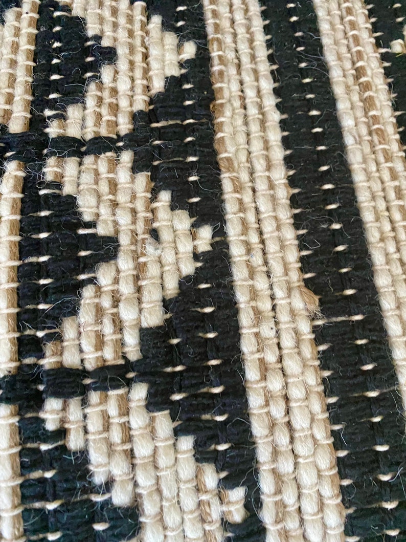Puede incluir: Primer plano de una alfombra tejida con un patr&oacute;n negro y crema. La alfombra est&aacute; hecha de fibras naturales y tiene una superficie texturizada.