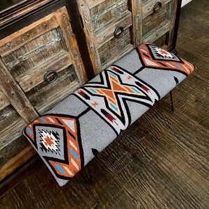 K&ouml;nnte beinhalten: Rechteckige Bank mit grau gemusterter Oberseite und schwarzen Haarnadelbeinen. Das Muster umfasst geometrische Formen in Orange, Schwarz, Wei&szlig; und Blau. Die Bank steht vor einem rustikalen Holzschrank.