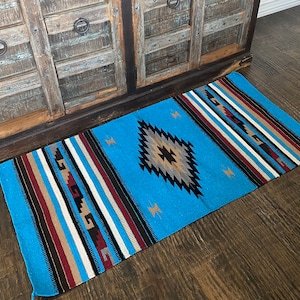 May include: A blue and brown patterned rug with a diamond design in the center. The rug is woven with stripes of different colors, including white, red, and black.