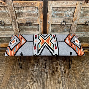 May include: A gray bench with a Southwestern-inspired design. The bench features a rectangular seat with a geometric pattern in orange, black, white, and blue. It has black metal hairpin legs, offering a modern touch to the rustic design.