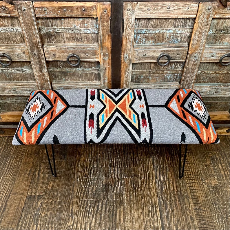 K&ouml;nnte beinhalten: Graue Bank mit einem Design im S&uuml;dwest-Stil. Die Bank hat eine rechteckige Sitzfl&auml;che mit einem geometrischen Muster in Orange, Schwarz, Wei&szlig; und Blau. Sie hat schwarze Haarnadelbeine aus Metall, die einen modernen Touch verleihen.