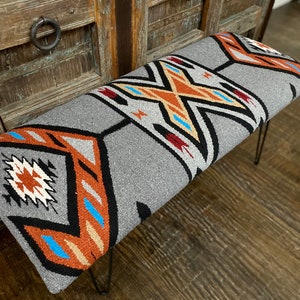 K&ouml;nnte beinhalten: Eine graue Bank mit einem Design im S&uuml;dweststil. Die Bank hat eine rechteckige Sitzfl&auml;che mit einem geometrischen Muster in Orange, Schwarz, Wei&szlig; und Blau. Die Beine sind d&uuml;nn, schwarz und aus Metall.