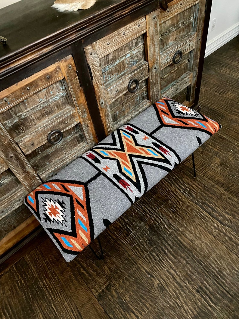 K&ouml;nnte beinhalten: Graue Bank mit einem Design im S&uuml;dwest-Stil. Der rechteckige Sitz hat ein geometrisches Muster in Orange, Schwarz, Wei&szlig; und Blau. Die Beine sind aus d&uuml;nnem, schwarzem Metall. Die Bank steht vor einem rustikalen Holzschrank. Ca. 120 cm lang.