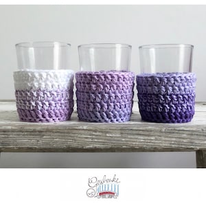 Könnte beinhalten: Drei Glasbecher mit gehäkelten Untersetzern in Ombre-Optik in Lila und Weiß. Die Untersetzer sind mit einem strukturierten Stich gearbeitet und haben einen Farbverlauf von Weiß nach Lila.