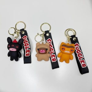 Peut inclure: Trois porte-clés Fuggler représentant des créatures de style dessin animé en noir, beige et orange. Chaque figurine a une large bouche avec des dents visibles, fixée à un anneau en métal doré et une sangle noire avec le mot FUGGLER en rouge.