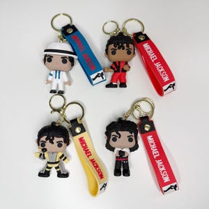 May include: A collection of Michael Jackson-themed keychains. Each keychain features a miniature figure of Michael Jackson in various iconic outfits. The keychains have gold-colored rings and straps with "MICHAEL JACKSON" printed on them.