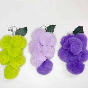 May include: Three fluffy grape-shaped keychains in shades of green, purple, and lavender. Each keychain features a cluster of soft, plush balls and a green leaf with red stitching. The keychains have silver metal rings.