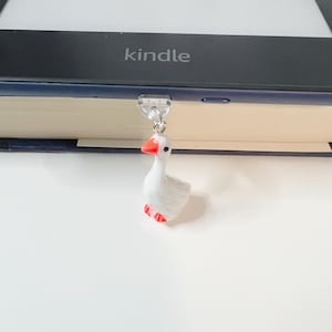 May include: A white goose bookmark with red beak and feet, hanging from a clear plastic bookmark holder. The bookmark is attached to a Kindle e-reader.