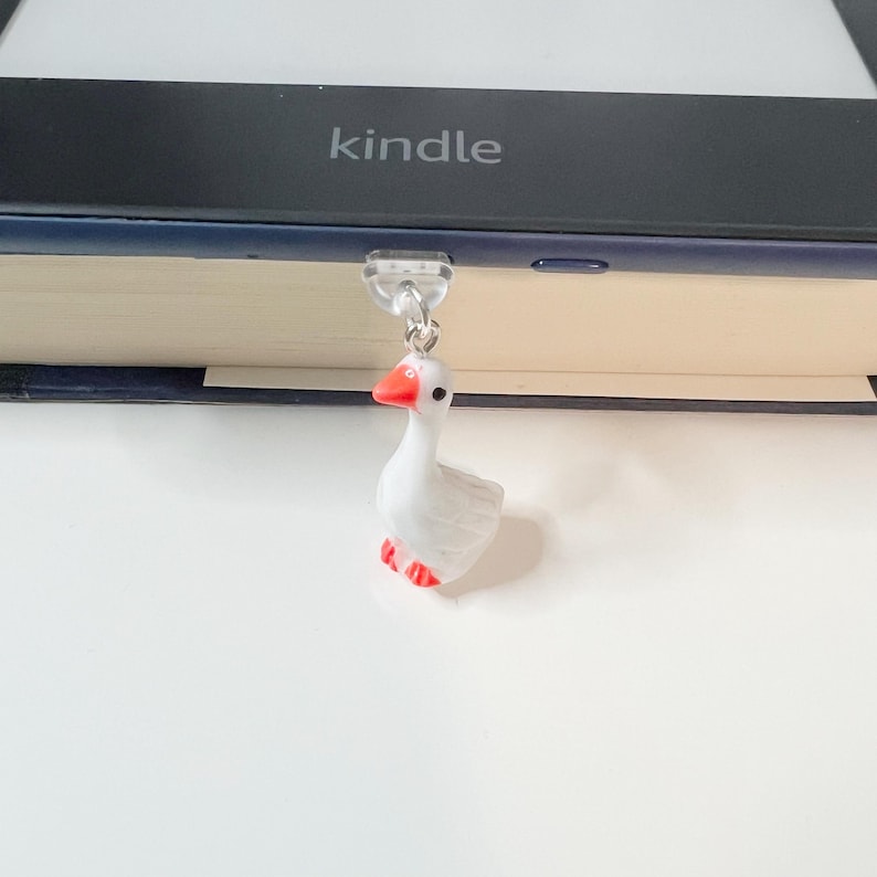 May include: A white goose bookmark with red beak and feet, hanging from a clear plastic bookmark holder. The bookmark is attached to a Kindle e-reader.