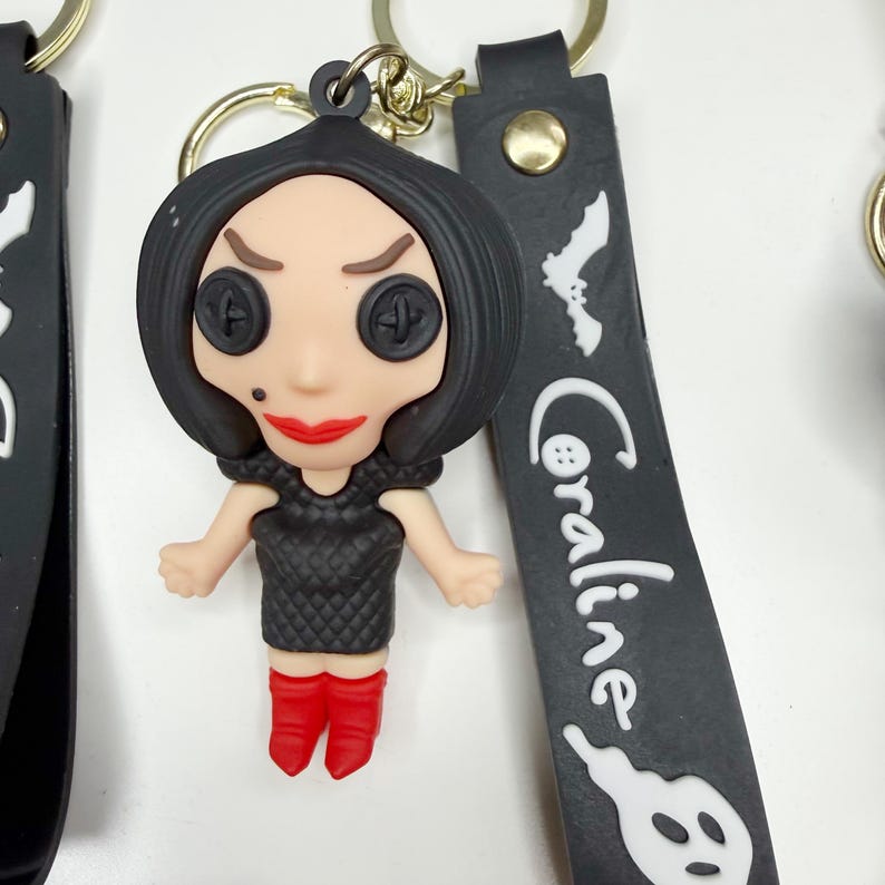 May include: A Coraline-themed keyring featuring a doll-like figure with button eyes, red lips, and red boots. The figure is wearing a black dress. The black strap has the word "Coraline" and a bat and ghost graphic.