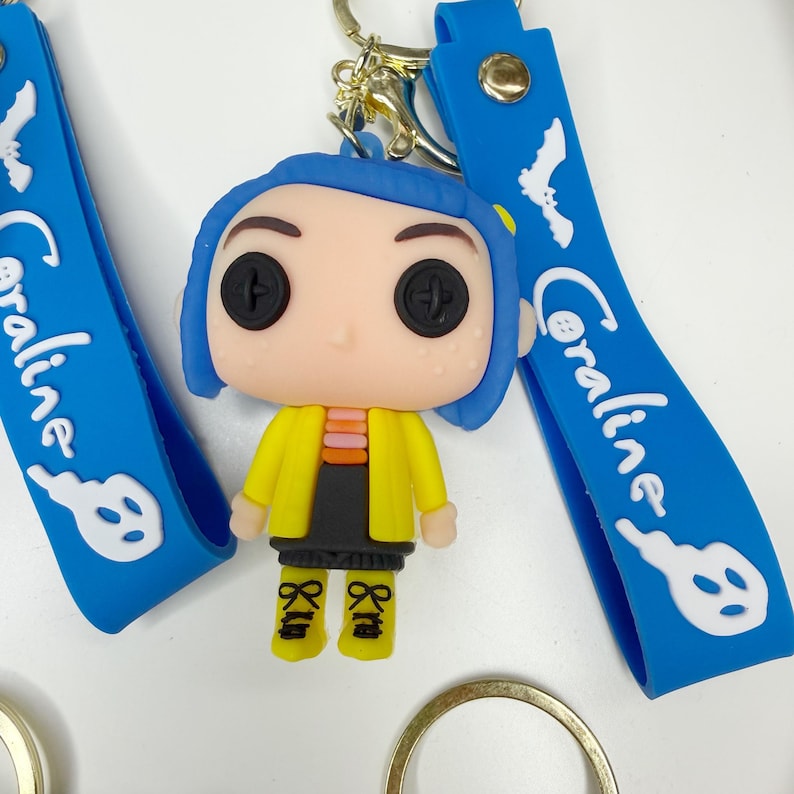 May include: A keychain featuring a figurine of a character with button eyes, blue hair, and a yellow jacket. The keychain includes a blue strap with the name "Coraline" and a keyhole design. The figurine is approximately 5 cm tall.