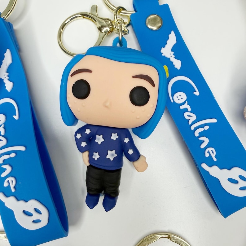 May include: A keychain with a figurine of a character with blue hair, wearing a blue sweater with white stars and black trousers. The keychain has a gold-coloured clasp and a blue strap with the name "Coraline" in white script.