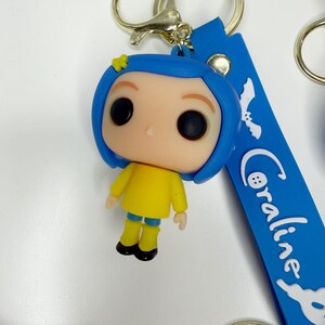 May include: A Coraline keychain featuring a stylized figure with blue hair, a yellow outfit, and large black eyes. The keychain has a blue strap with the name "Coraline" in white script and a gold-coloured keyring.