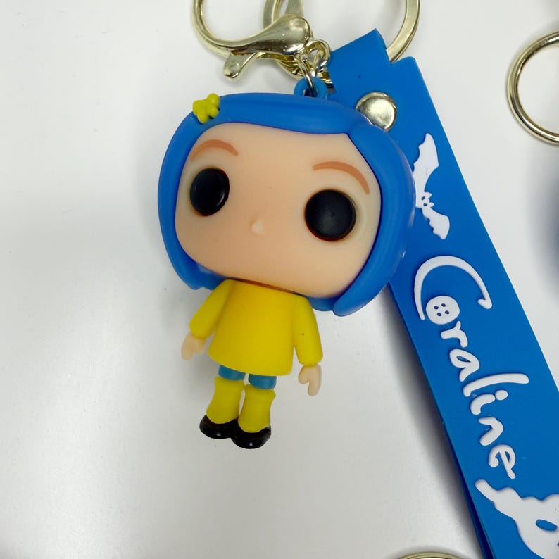 May include: A Coraline keychain featuring a stylized figure with blue hair, a yellow outfit, and large black eyes. The keychain has a blue strap with the name "Coraline" in white script and a gold-coloured keyring.